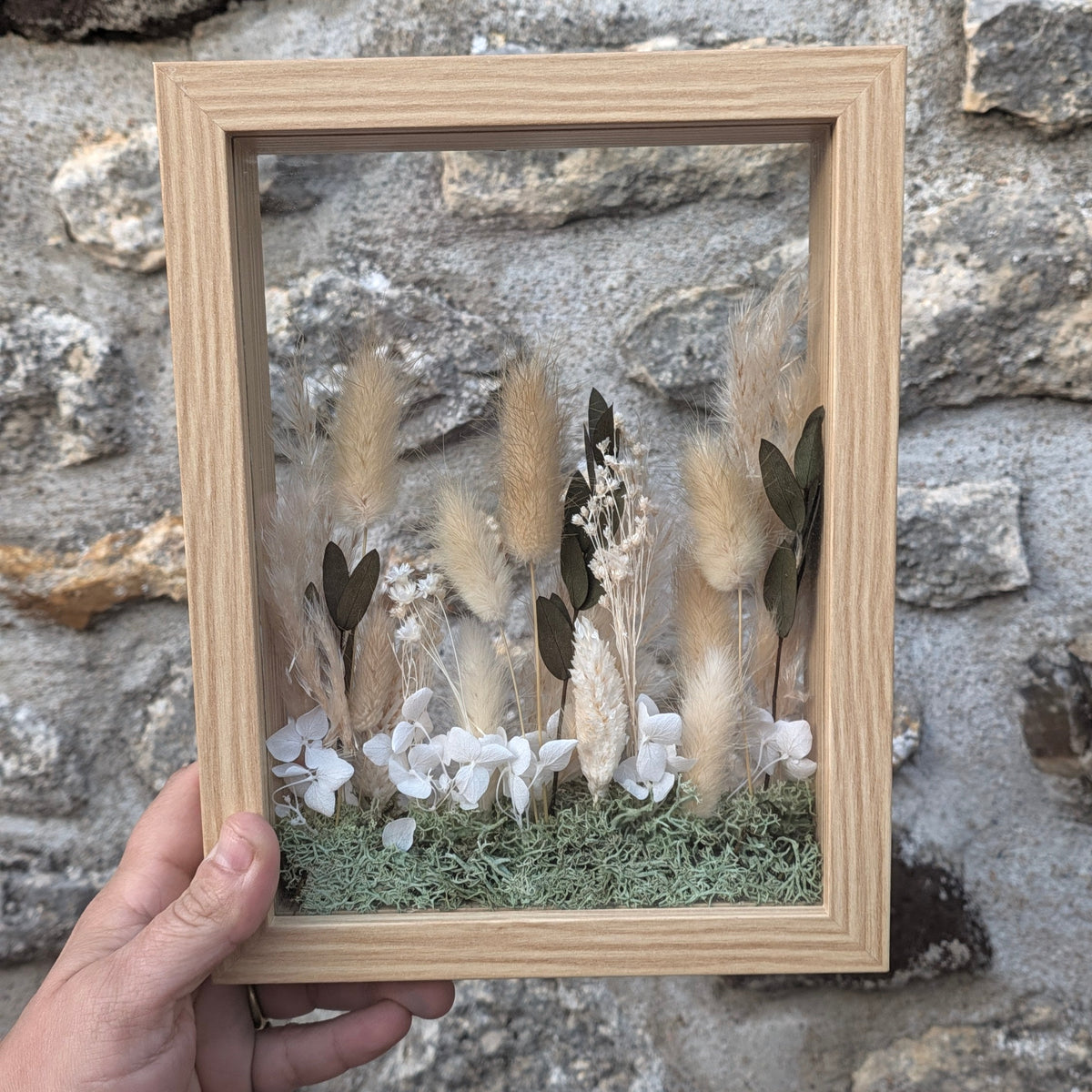Cadre La Clairière tenu en main pour visualiser son format, petit paysage végétal en volume réalisé artisanalement avec des fleurs séchées.