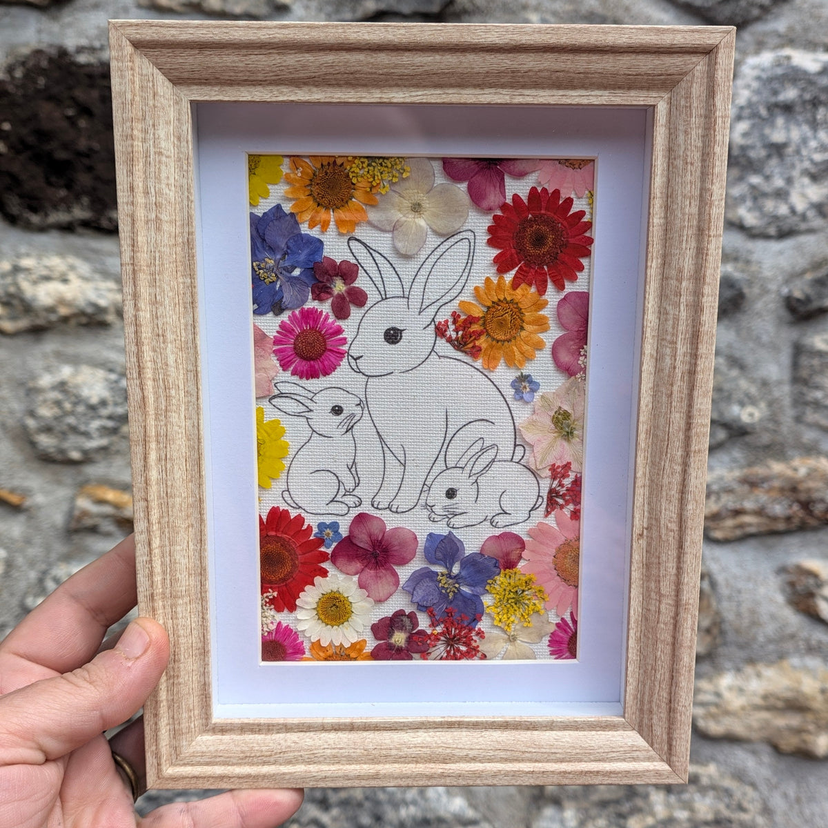 Cadre fleuri La famille de Max tenu en main pour visualiser son format, avec illustration d’une famille de lapins entourée de véritables fleurs pressées colorées.