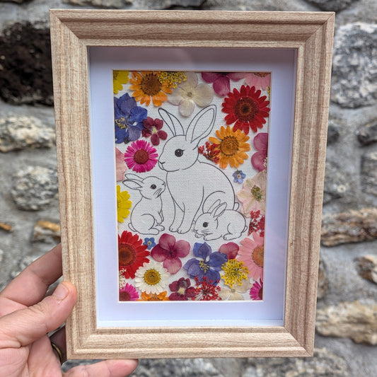 Cadre fleuri La famille de Max tenu en main pour visualiser son format, avec illustration d’une famille de lapins entourée de véritables fleurs pressées colorées.