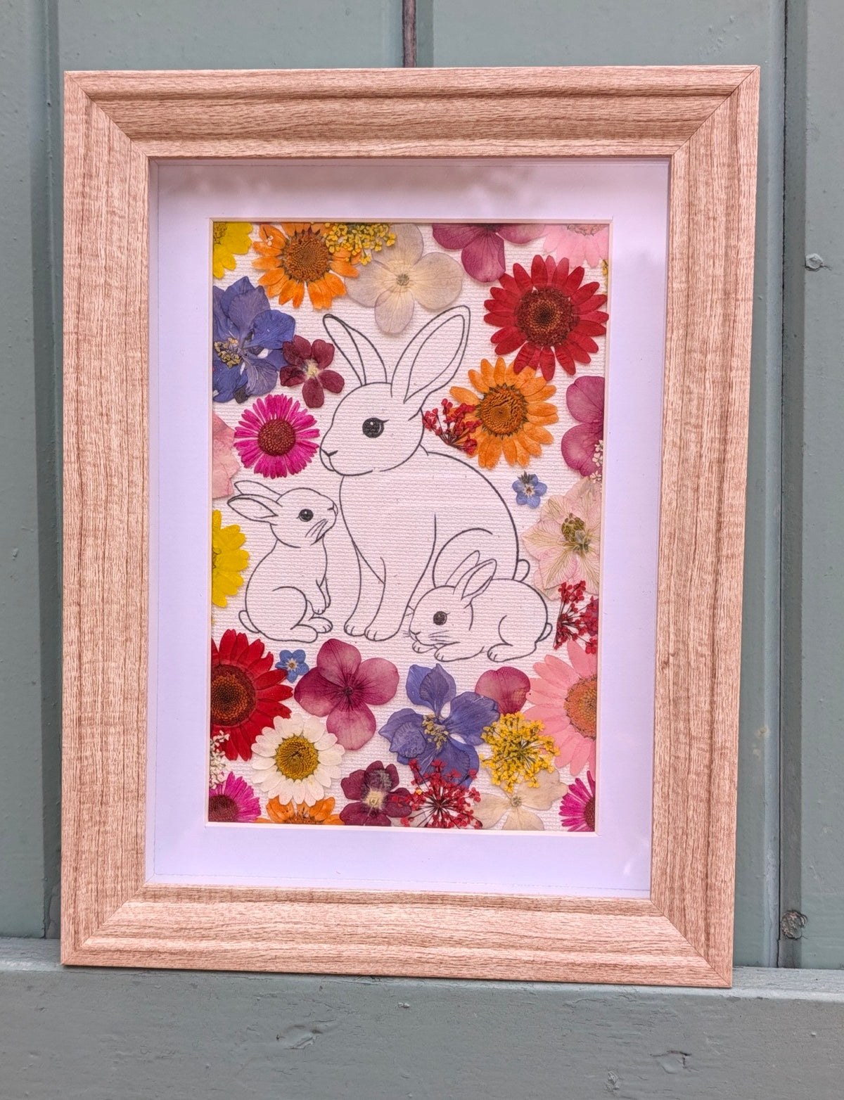 Cadre La famille de Max de la collection Faunéa, illustration d’une maman lapin et ses deux petits entourés de véritables fleurs séchées sous presse.